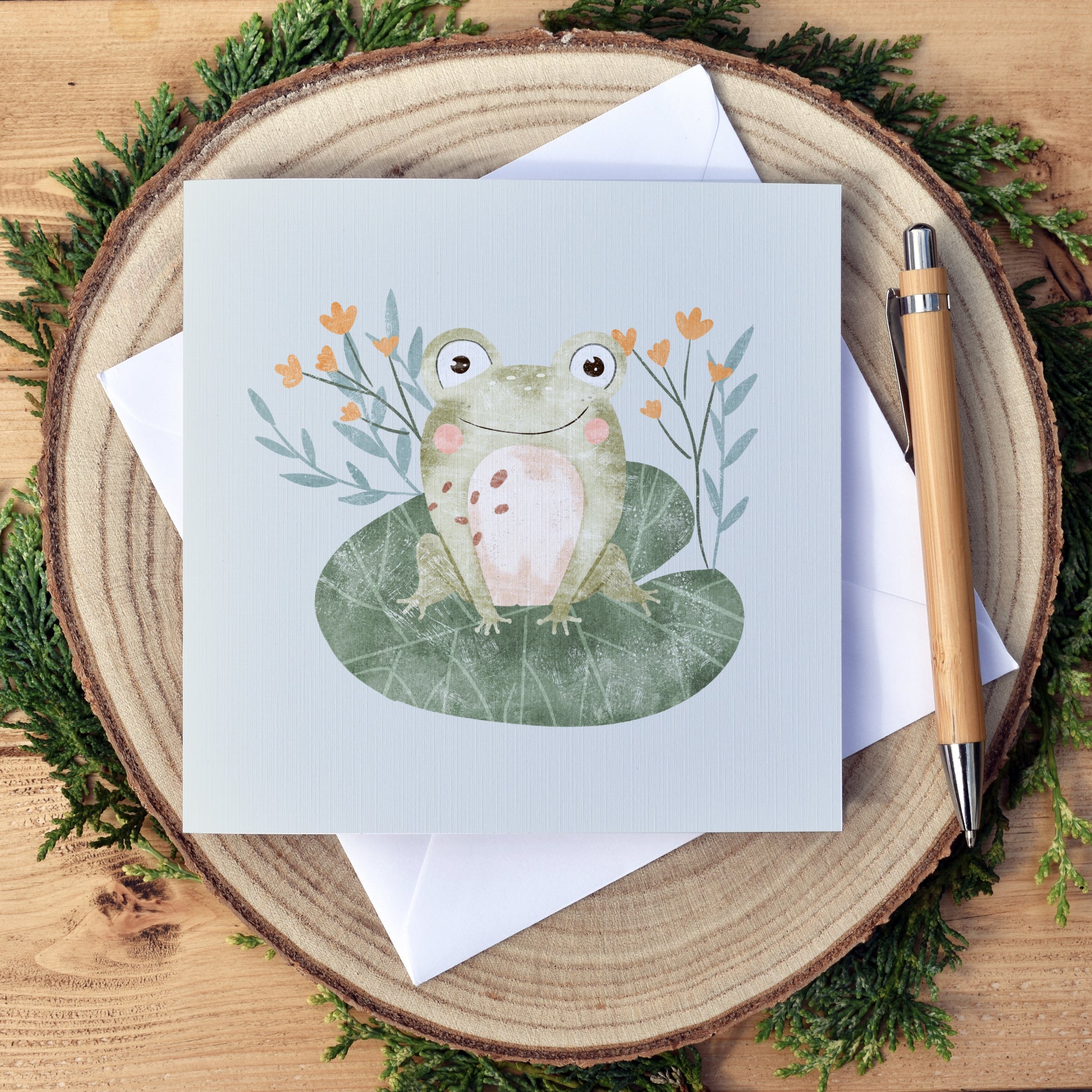 Cute frog sitting on a Lilypad, with some simple greenery and yellow flowers in the background. The card can be personalised with the child's name that sits under the frog illustration. The greeting at the top reads happy birthday. A plain, no text version is also available. The card once folded is approximately 6x6 inches and comes with a white envelope.