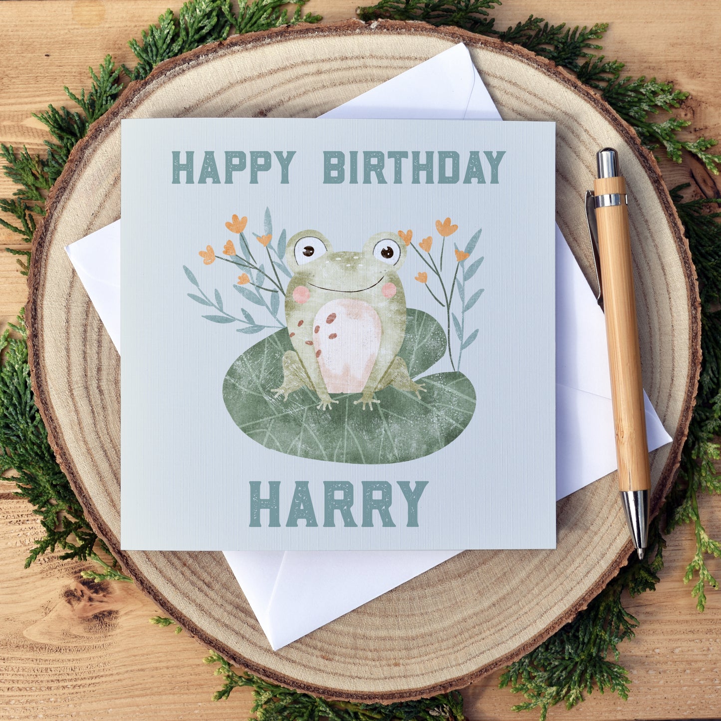 Cute frog sitting on a Lilypad, with some simple greenery and yellow flowers in the background. The card can be personalised with the child's name that sits under the frog illustration. The greeting at the top reads happy birthday. A plain, no text version is also available.  The card once folded is approximately 6x6 inches and comes with a white envelope. 