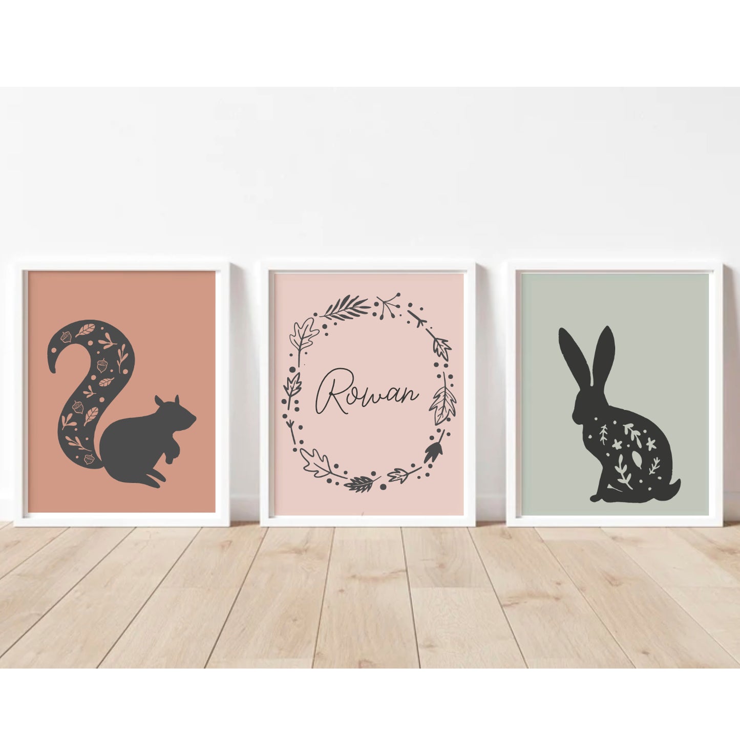 Trio of woodland art prints, squirrel silhouette with leaf and acorn pattern in the tail, on a terracotta boho shade of orange. A simple woodland leaf wreath with personalisation in the centre, on a dusky pink background, Rabbit silhouette with leaf pattern, on a sage green background. The name can be customised to any name or saying  